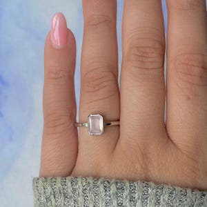 May include: A silver ring with a rectangular, light pink gemstone. The ring is set in a simple, minimalist style. The gemstone is the focal point, showcasing its soft color and shape. The ring is on a finger.