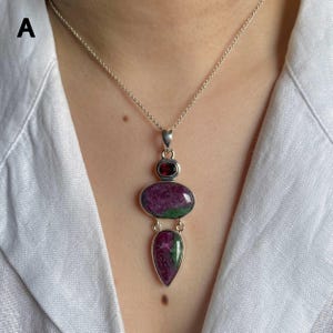 May include: A silver pendant necklace featuring a small round dark red gemstone at the top, a larger oval-shaped ruby and zoisite stone in the center, and a teardrop-shaped ruby and zoisite stone at the bottom, all set in polished silver metal.