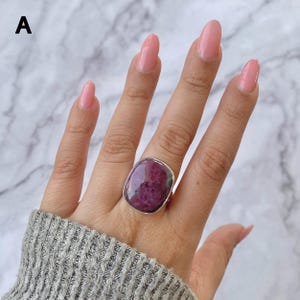 May include: A silver ring featuring a large, smooth, oval-shaped magenta and dark purple gemstone set in a simple metal bezel. The stone displays natural mottled color variations throughout its surface, and the ring is worn on a finger.