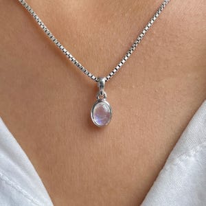 May include: A silver necklace with a small, oval pendant. The pendant holds a light-coloured, iridescent stone, possibly a moonstone. The chain is a delicate box chain style. The necklace is shown against a neutral background.