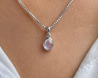 Könnte beinhalten: Eine silberne Halskette mit einem kleinen, ovalen Anhänger. Der Anhänger hat einen hellfarbigen, irisierenden Stein, möglicherweise einen Mondstein. Die Kette ist eine filigrane Boxkette. Die Halskette wird vor einem neutralen Hintergrund gezeigt.