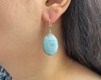 May include: An oval-shaped Larimar gemstone earring set in a polished silver bezel with a lever-back closure. The stone features a marbled pattern of light blue, turquoise, and white tones, creating a smooth, rounded surface within the metal frame.