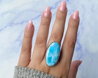 May include: A large oval ring featuring a polished blue and white Larimar gemstone set in a smooth silver bezel. The stone displays a distinct marbled pattern with varying shades of sky blue and white, securely mounted on a simple silver band.