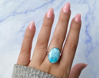 May include: A silver ring featuring a large, oval-shaped, sky-blue Larimar gemstone with white marbled patterns. The stone is set in a smooth, polished metal bezel, and the band is a simple, thin silver wire that encircles the finger.