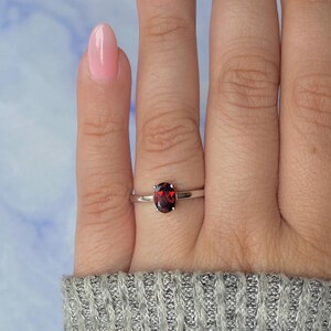 May include: A silver ring with a dark red oval gemstone. The ring is on a finger, and the background is a light blue and white color.