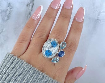 Könnte beinhalten: Ein silberner Ring mit einem ovalen K2-Stein mit blauen und weißen Mustern, akzentuiert durch drei blaue Edelsteine. Der Ring wird an einer Hand mit einem grauen Pullover getragen.