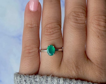 May include: A silver ring featuring a vibrant oval-cut green gemstone. The ring is set on a slender band, showcasing the polished gemstone. The ring is displayed on a finger, highlighting the jewelry's design and color.