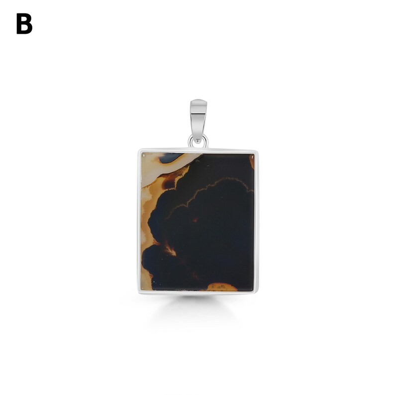 May include: A rectangular pendant featuring a polished stone with a dark black center and cream-colored edges, set in a silver-toned metal frame with a small bail at the top. The letter B is visible in the upper left corner of the image.