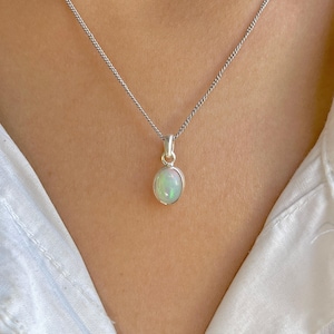 May include: A silver necklace featuring an oval opal pendant. The opal displays a milky white hue with iridescent green and blue flashes. The pendant is set in a silver bezel and suspended from a fine silver chain. The necklace is shown against a white backdrop.