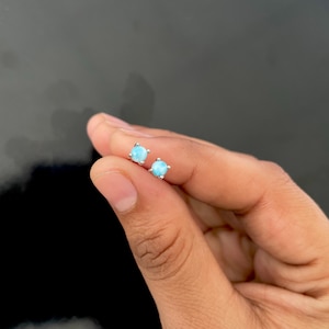 May include: A pair of light blue stud earrings with a round, polished gemstone set in a silver metal. The earrings are held in a person's hand against a dark background. The earrings are small and delicate.