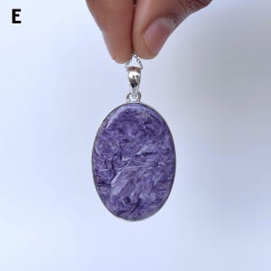 Op de afbeelding: Een ovaalvormige hanger met een gepolijste paarse charoiet edelsteen met wervelende witte en donkerpaarse patronen, gezet in een eenvoudige zilverkleurige metalen rand met een klein oogje aan de bovenkant. De letter E is linksboven zichtbaar.