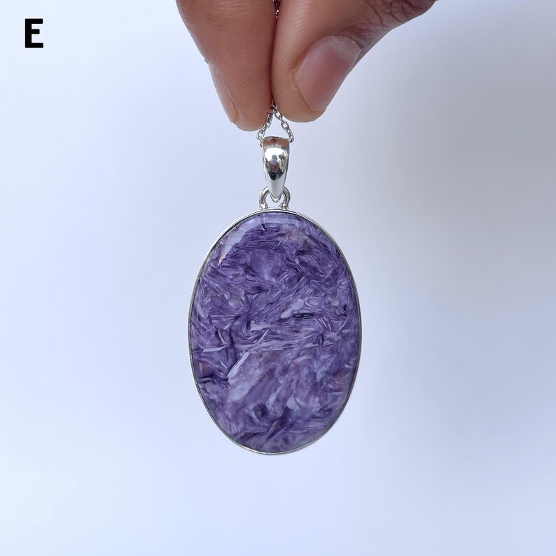 Op de afbeelding: Een ovaalvormige hanger met een gepolijste paarse charoiet edelsteen met wervelende witte en donkerpaarse patronen, gezet in een eenvoudige zilverkleurige metalen rand met een klein oogje aan de bovenkant. De letter E is linksboven zichtbaar.
