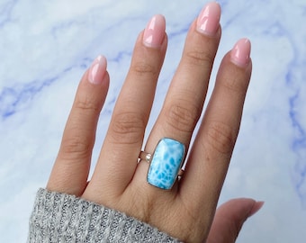 May include: A silver ring featuring a large, rectangular, light blue Larimar gemstone with white marbled patterns. The stone is set in a smooth metal bezel, and the band is a simple, polished silver metal. The letter A is visible in the top corner.