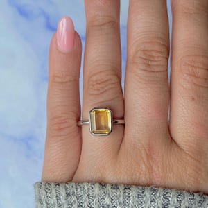 May include: A silver ring with a rectangular, golden-yellow gemstone. The ring has a simple, modern design, highlighting the stone's vibrant colour. The ring is a piece of jewellery.