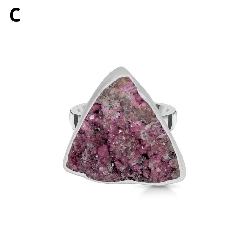 May include: A silver ring featuring a triangular, rough-cut gemstone. The stone is a mix of pink and gray hues, set within a polished silver band. The ring's design highlights the natural texture and color variations of the stone.