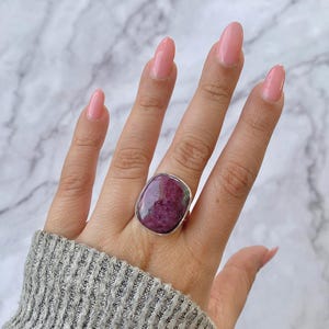 May include: A silver ring with a large, rectangular, polished ruby gemstone. The ruby is a deep red and purple colour with green inclusions. The ring is set on a silver band and is shown on a hand.