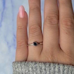 Peut inclure: Une bague en argent avec une petite pierre noire ronde. La bague a un design simple, avec un fin anneau et un sertissage à lunette pour la pierre sombre. La bague est portée au doigt.