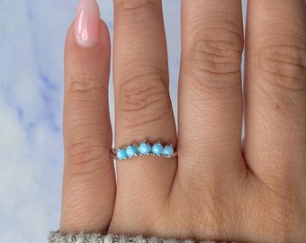 May include: A silver ring featuring five small, round, light blue gemstones set in a row. The ring is shown on a person's finger, with a light blue and white background.