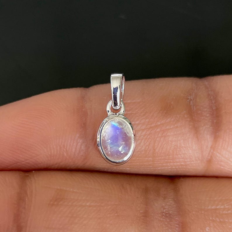 May include: A small, oval pendant with a silver-coloured bail. The pendant features a polished, iridescent moonstone with blue and white hues. The gemstone is set in a silver bezel.