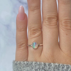May include: A silver ring with an oval opal gemstone. The opal displays a play of color with hues of green, blue, and yellow. The ring is set on a slender band and is shown on a hand.