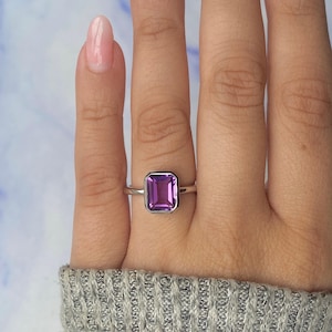 May include: A silver ring with a rectangular, deep purple gemstone. The gemstone is set in a bezel setting. The ring is shown on a hand.
