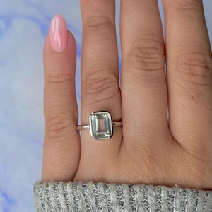 May include: A silver ring featuring a rectangular, pale green gemstone. The ring has a minimalist design with a slender band and a bezel setting. The gemstone is transparent and displays a delicate, light green colour.