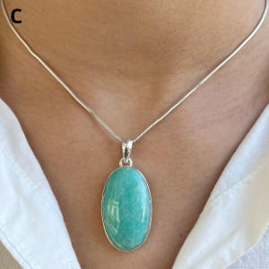May include: A silver necklace featuring a smooth, oval-shaped, light turquoise-colored gemstone pendant. The pendant is set in a simple silver frame and hangs from a thin, flexible silver snake chain. The letter C is visible in the top left corner.
