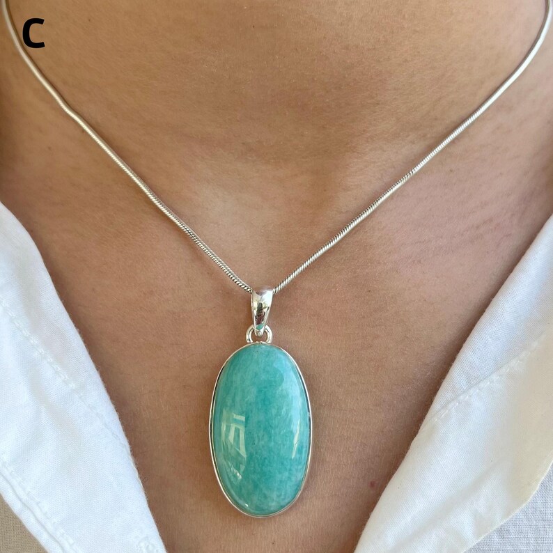 May include: A silver necklace featuring a smooth, oval-shaped, light turquoise-colored gemstone pendant. The pendant is set in a simple silver frame and hangs from a thin, flexible silver snake chain. The letter C is visible in the top left corner.