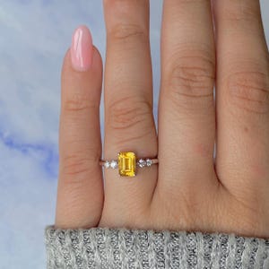 May include: A silver ring featuring a rectangular, yellow gemstone as the focal point. The ring is accented with small, round, clear gemstones on either side of the main stone. The ring is set on a finger with a light pink nail polish.