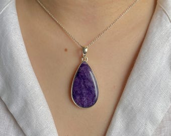 May include: A teardrop-shaped purple charoite pendant set in a polished silver bezel, suspended from a delicate silver chain. The stone features a swirling, marbled texture with deep violet and white inclusions, held by a simple silver bail.