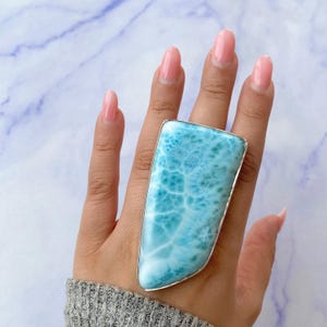 May include: A large, rectangular ring featuring a light blue larimar stone with white veining, set in a silver-colored metal. The ring is displayed on a hand with manicured nails, against a marble-patterned background.