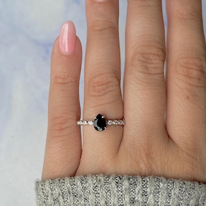 May include: A silver ring with a pear-shaped black gemstone at its centre. The band is embellished with small, clear gemstones. The ring is shown on a hand with painted nails, set against a light blue and white backdrop.