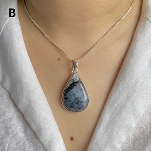 May include: A teardrop-shaped pendant featuring a translucent white stone with dark, fern-like dendritic inclusions, encased in a smooth silver setting. The pendant hangs from a delicate silver box chain necklace, resting against a white fabric collar.