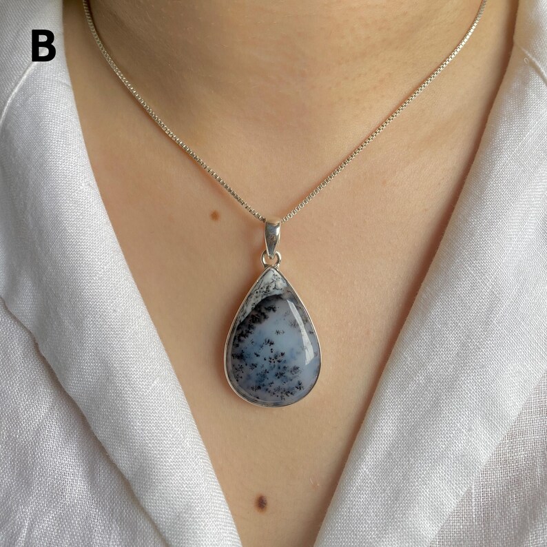 May include: A teardrop-shaped pendant featuring a translucent white stone with dark, fern-like dendritic inclusions, encased in a smooth silver setting. The pendant hangs from a delicate silver box chain necklace, resting against a white fabric collar.