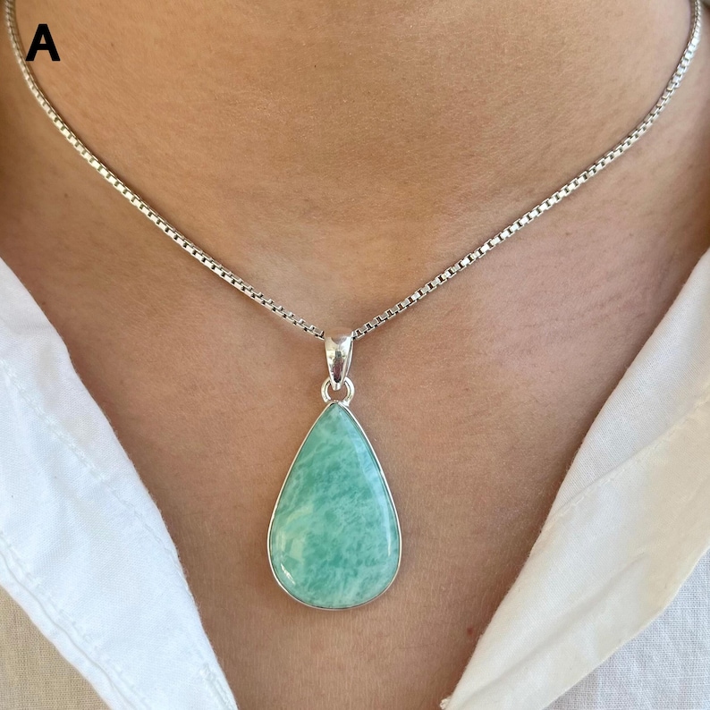 May include: A silver box chain necklace featuring a teardrop-shaped pendant with a smooth, light green stone. The pendant is set in a polished silver frame and hangs from a delicate, square-linked silver chain resting against the collarbone.