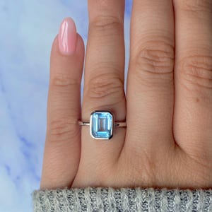 May include: A silver ring with a rectangular, light blue gemstone. The gemstone is set in a bezel setting. The ring is shown on a hand with pink nail polish.