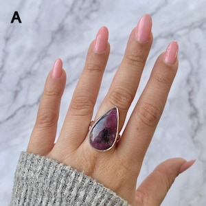 Puede incluir: Un anillo de plata con una gran piedra preciosa pulida en forma de lágrima, con un patrón moteado en magenta intenso y negro. La piedra está engastada en un bisel de plata liso y sencillo, y la banda del anillo tiene un acabado de plata pulida.