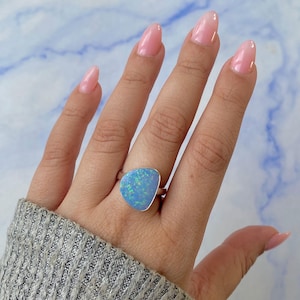 Può includere: Un anello d'argento con una grande pietra di opale azzurro chiaro dalla forma irregolare. L'opale mostra giochi di colore con sfumature di verde e blu. L'anello è indossato su una mano con smalto rosa e un maglione grigio.