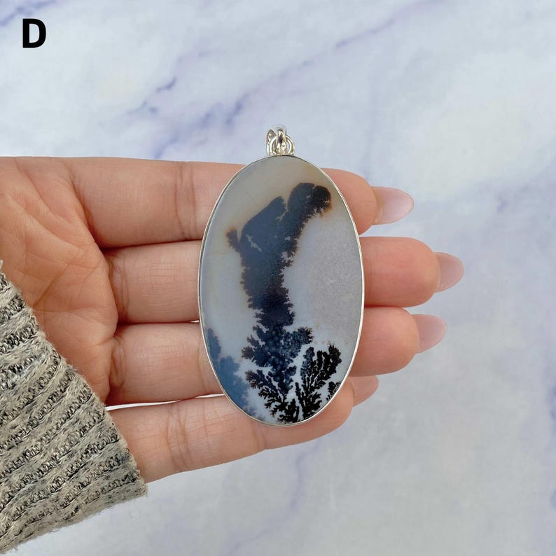 May include: An oval-shaped dendritic agate pendant set in a silver-toned metal frame. The stone features a translucent, milky white base with dark, fern-like inclusions that branch out across the surface. A small silver bail is attached at the top.