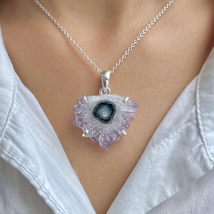 Puede incluir: Collar de plata con colgante de rodaja de geoda. La geoda tiene un centro azul oscuro y blanco, rodeado de una formación de cristales de color púrpura claro. El collar se muestra sobre una camisa blanca.