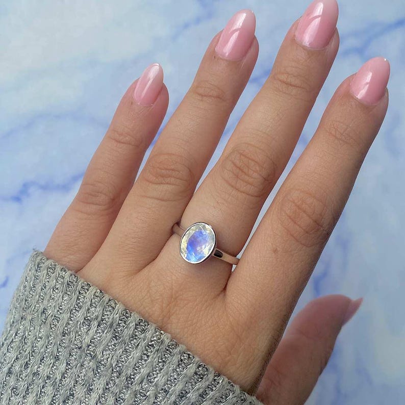 May include: A silver ring featuring an oval-shaped, iridescent moonstone. The gemstone displays a play of blue and white colors. The ring is set on a slender band. The background is a light blue and white marble pattern.