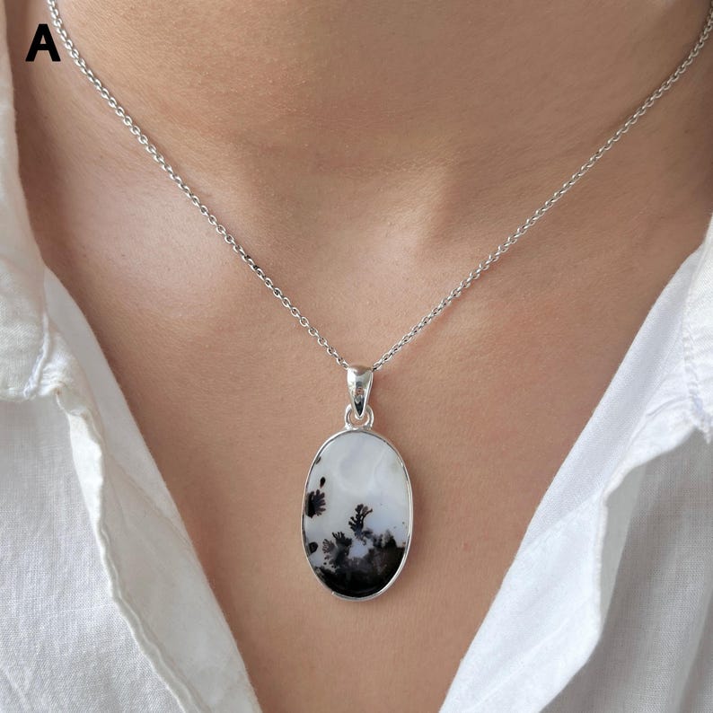 Puede incluir: Un colgante de forma ovalada con una piedra blanca que presenta inclusiones dendr&iacute;ticas negras similares a helechos, engastado en un marco de metal plateado pulido. El colgante cuelga de una delicada cadena plateada sobre una camisa blanca.