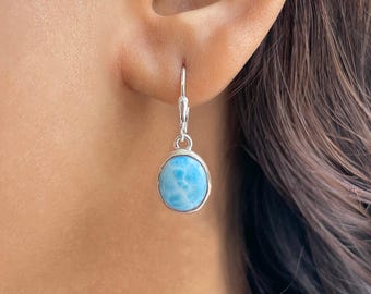 Könnte beinhalten: Ein silberner Ohrhänger mit einem glatten, ovalen, hellblauen Larimar-Edelstein mit weißen Marmorierungen. Der Stein ist in einer polierten Silberfassung eingefasst und hängt an einem einfachen silbernen Klappbügel.