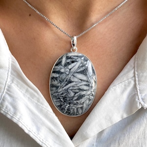 May include: Oval pendant necklace with a silver chain. The pendant features a black and white stone with a unique, abstract pattern. The necklace is worn against a white shirt.
