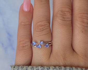 May include: A silver ring with five light blue gemstones. The gemstones are in a variety of shapes, including pear and round cuts. The ring is on a finger with a light pink nail polish.