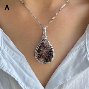 May include: A teardrop-shaped pendant featuring a dark brown, fibrous stone set in a silver frame with a twisted rope border. The pendant hangs from a silver chain with a classic link design, resting against a white collared shirt.