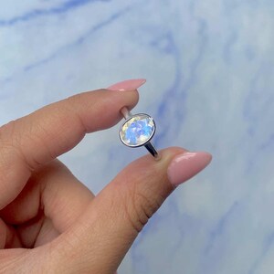 May include: A silver ring featuring an oval-shaped, iridescent gemstone. The gemstone displays a mix of blue, white, and yellow hues. The ring is held against a light blue and white marbled background.