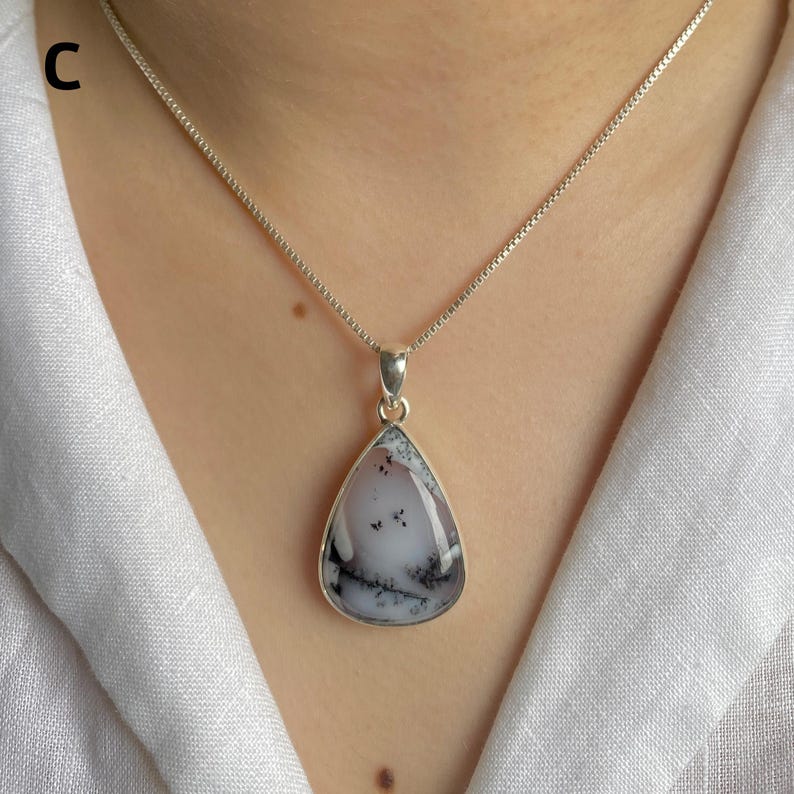 May include: A teardrop-shaped pendant featuring a milky white stone with dark, fern-like dendritic inclusions, encased in a polished silver setting. The pendant hangs from a delicate silver box chain necklace against a white fabric background.