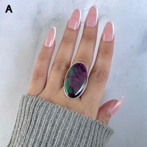 May include: A ring featuring a large, oval-shaped cabochon stone with a polished surface displaying deep magenta and vibrant green color patterns. The stone is set in a smooth, plain silver-toned metal bezel that encircles the entire gemstone.