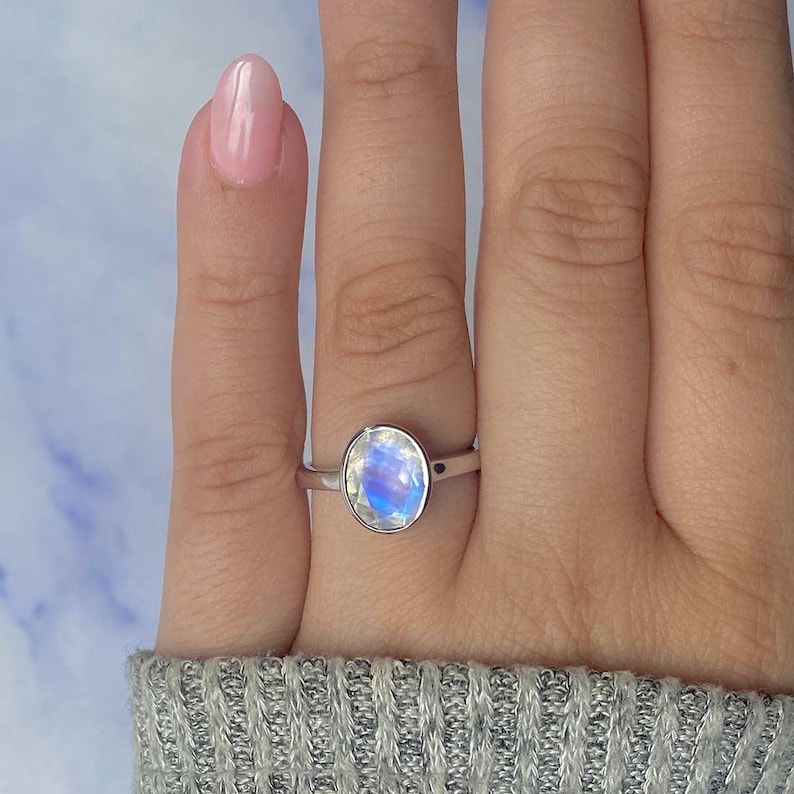 May include: A silver ring with an oval, iridescent moonstone. The moonstone displays a mix of blue, white, and yellow hues. The ring is set on a finger, with a light pink nail polish visible.